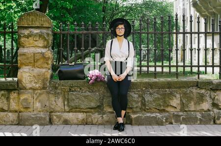 A beautiful girl with hat on her head and in glasses sita on the stone fence and looking to the sides. Beautiful summer day. Stock Photo