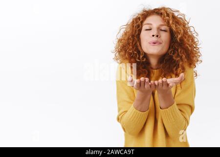 Dreamy, lovely tender and romantic redhead curly woman, wear yellow sweater, send her passionate air kiss at camera, holding palms near folded lips Stock Photo