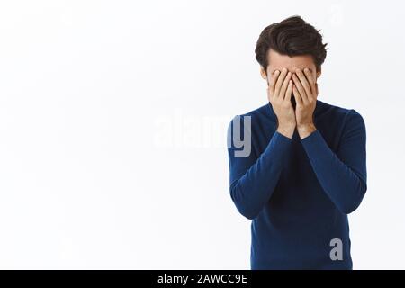 Distressed, tired businessman in blue high neck sweater, hiding face, facepalm, holding hands on head and sighing, feeling troubled, exhausted as Stock Photo