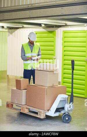 Young man in working clothing making notes in document while standing near the cart with boxes in storage room Stock Photo