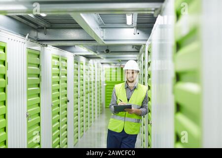 Portrait of young man in working clothing looking at camera while making notes in warehouse Stock Photo