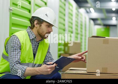 Young man in work helmet sitting with document and pointing at cardboard box he checking the address before delivering Stock Photo