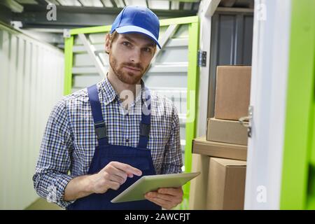 Portrait of young bearded manual worker in overalls looking at camera while using digital tablet in warehouse Stock Photo