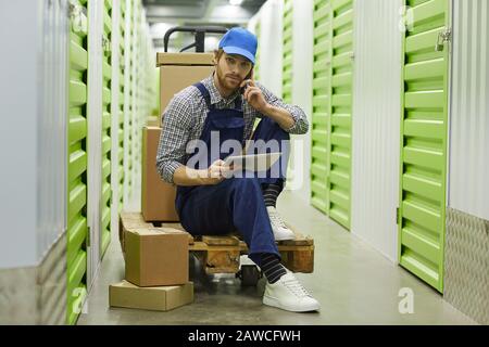 Young worker in overalls sitting on cart with tablet pc and discussing work with somebody on mobile phone in storage room Stock Photo
