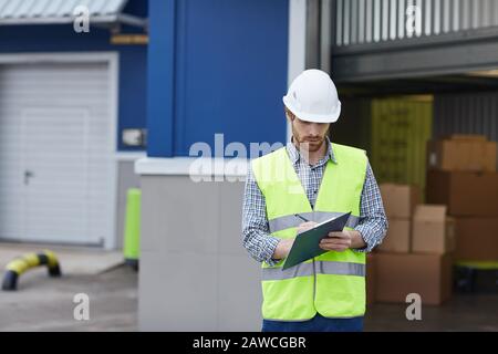 Young man in protective workwear standing and making notes in document while delivering load he working outdoors Stock Photo