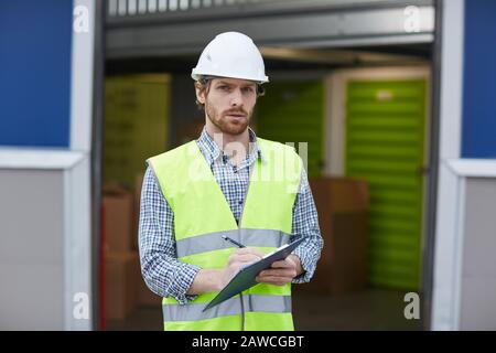 Portrait of young worker in work helmet and in reflective vest looking at camera while making notes and making delivery outdoors Stock Photo