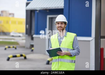 Portrait of bearded young man in protective workwear smiling at camera while standing outdoors with document Stock Photo