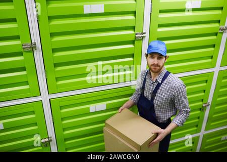 Portrait of young man in cap and in overalls holding box and looking at camera while standing near the green boxes in stockroom Stock Photo