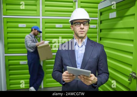 Portrait of confident businessman in suit and in work helmet using tablet pc and looking at camera with worker working in the background Stock Photo