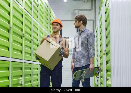 Young manual worker in work helmet carrying box and talking with trainee during their work in warehouse Stock Photo