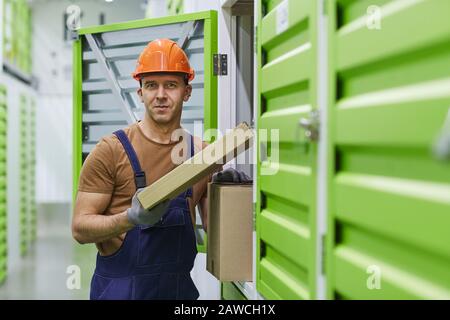 Portrait of young worker in work helmet and in overalls looking at camera while putting parcels into the boxes in warehouse Stock Photo
