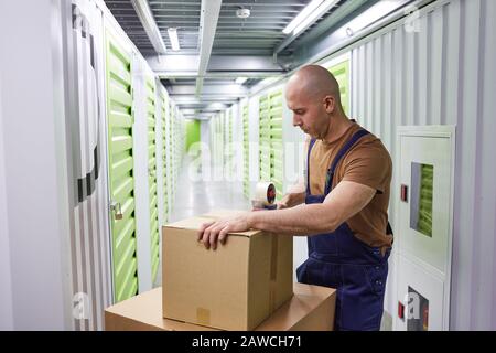 Young man in overalls sealing the parcels with tape while standing in storage room Stock Photo