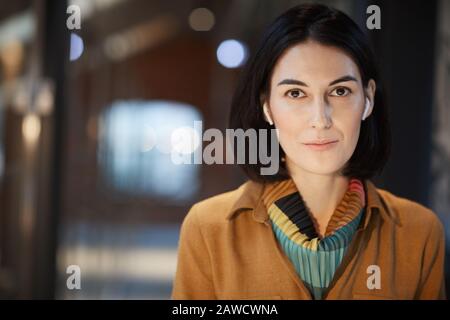 Portrait of young businesswoman with dark hair and in wireless headphones looking at camera while working at office Stock Photo