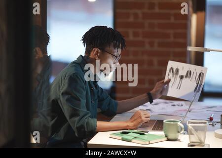 Serious African designer in eyeglasses concentrating on his work on laptop and examining sketches of clothes at his workplace at office Stock Photo