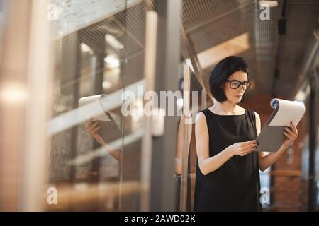 Serious young businesswoman in eyeglasses and in elegant black dress standing and reading a document Stock Photo