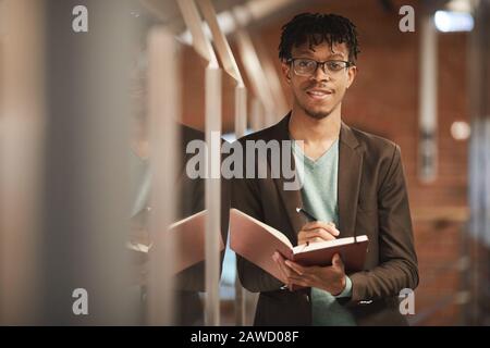 Portrait of African young businessman in eyeglasses smiling at camera while standing with note pad and planning his work Stock Photo