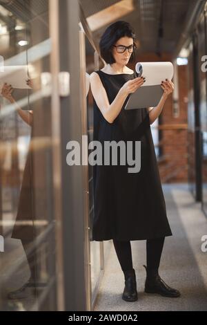 Young businesswoman in black dress examining business document while standing at office corridor Stock Photo