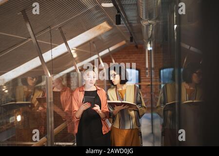 Young businesswoman using digital tablet and talking to her colleague with note pad while they walking along the office corridor Stock Photo