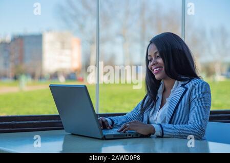 African-American woman using a laptop in an office with a window overlooking a park Stock Photo