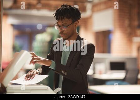 Young African businessman in eyeglasses standing near the printer and making copy of documents at office Stock Photo
