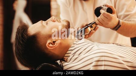 Barber shaves his beard to hipster man in barbershop. Toned photo, vintage style. Stock Photo