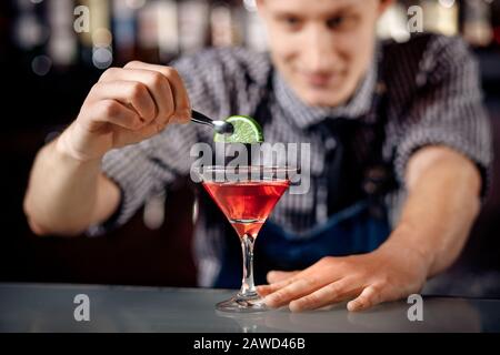 Expert bartender making cocktail with shaker in martini glass, red. Dark background. Stock Photo