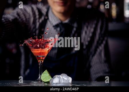 Barman is preparing red cocktail in martini glass. Splashes, dark background. Stock Photo