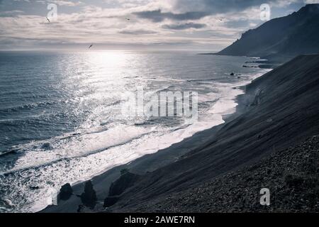 view of Icelandic rocky coast on sunny day with sun shining through clouds and birds flying over sea Stock Photo