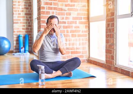 Middle age handsome sportman sitting on mat doing stretching yoga exercise at gym rubbing eyes for fatigue and headache, sleepy and tired expression. Stock Photo