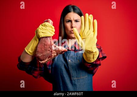 Young butcher woman holding fresh raw beef meat stake over red background with open hand doing stop sign with serious and confident expression, defens Stock Photo