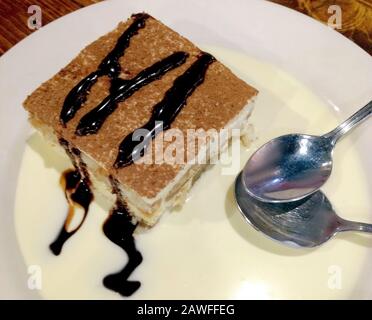 Delicious slice of tiramisu on a vanilla cream sauce, drizzled with chocolate and served with two spoons for sharing Stock Photo