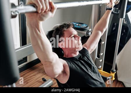 Determined young bodybuilder lifting barbell when sitting on gym bench Stock Photo