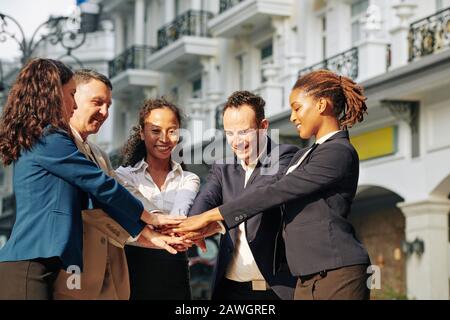 Positive successful business executives standing in the street in sunset sun and stacking hands to celebrate success after closing deal Stock Photo