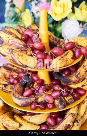 Close-up view of overripe spotted bananas and red grapes placed on a plastic tower tray, with colorful plastic flowers seen in the background Stock Photo
