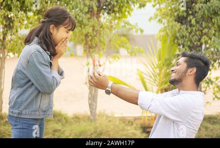 Concept of teenage love or affection - young man proposing to smiling excited girlfriend standing on knee with red rose on valentines day Stock Photo