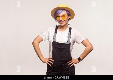 Style, fashion. Portrait of bright fashionably dressed hipster girl with violet short hair in sunglasses and hat holding hands on hips, looking confid Stock Photo