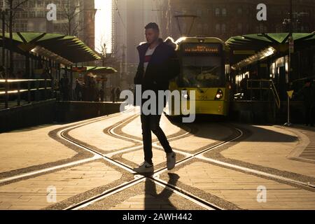 A man walks across the on-street track at the Metrolink tram stop St Peter's Square Manchester with a yellow tram in the background. Stock Photo