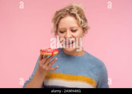 Tasty dessert. Portrait of woman with short curly hair in sweater holding doughnut, looking with desire at donut and licking her lips, wants to eat sw Stock Photo
