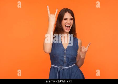 Hard rock lover. Delighted crazy brunette woman in denim dress making devil horns gesture with fingers up and roaring yelling, rock and roll dance mov Stock Photo