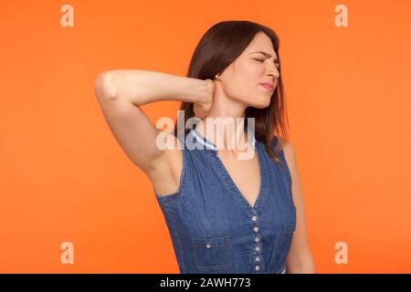 Hurting neck. Tired unhealthy brunette woman in denim dress standing massaging neck and grimacing in pain, suffering muscle tension, injured backbone. Stock Photo