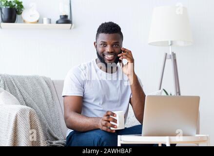 Positive african guy working on laptop and calling on phone Stock Photo