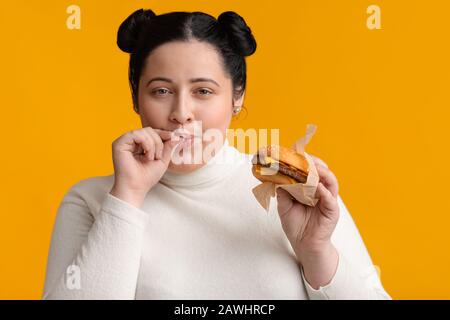 Young obese girl eating burger and licking fingers, enjoying fast food Stock Photo