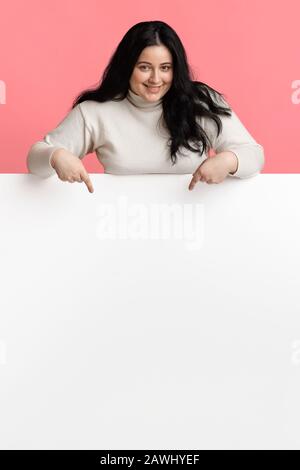 Young overweight woman pointing at white blank advertisement board Stock Photo