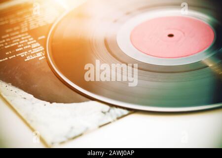 A vintage musical black vinyl record lies on the paper cover of a music album, illuminated by sunlight. Stock Photo