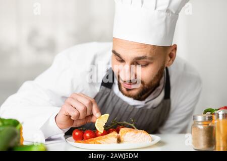 Professional Chef Plating Roasted Salmon Standing In Kitchen Stock Photo
