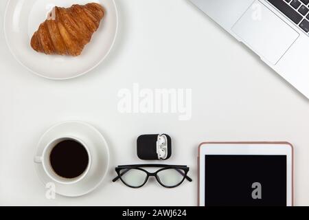 Business breakfast concept. Modern unisex working space, top view. Laptop, coffee, tablet, croissant, glasses, headphones in case on white background, Stock Photo