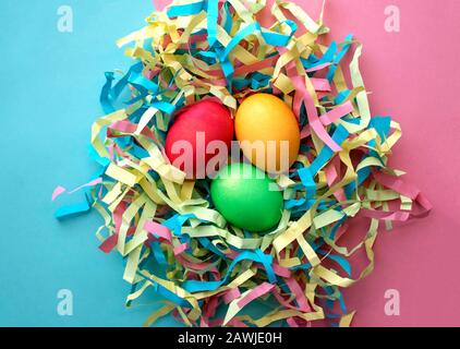 Three dyed Easter eggs in a colorful nest on mixed pink and blue background. Top view. Symbol of main Christian feast celebration. Stock Photo