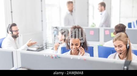 Call center worker accompanied by her team. Smiling customer support operator at work. Young employee working with a headset. Stock Photo