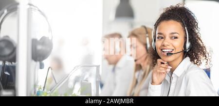 Call center worker accompanied by her team. Smiling customer support operator at work. Young employee working with a headset. Stock Photo