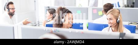 Call center worker accompanied by her team. Smiling customer support operator at work. Young employee working with a headset. Stock Photo